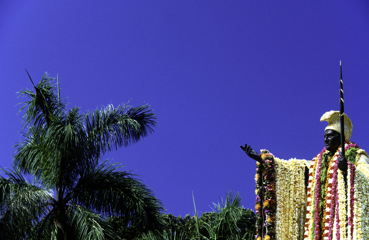 King Kamehameha's statue adorned with vibrant flower lei, set against a clear blue sky and surrounded by palm trees.