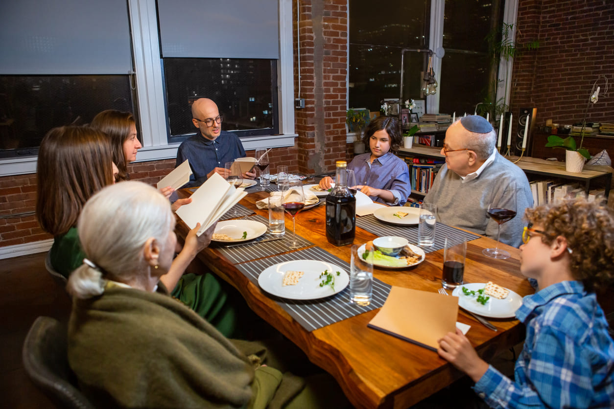 A family gathers around the table, reading from Passover texts and sharing a meal with wine and matzah to honor the holiday.