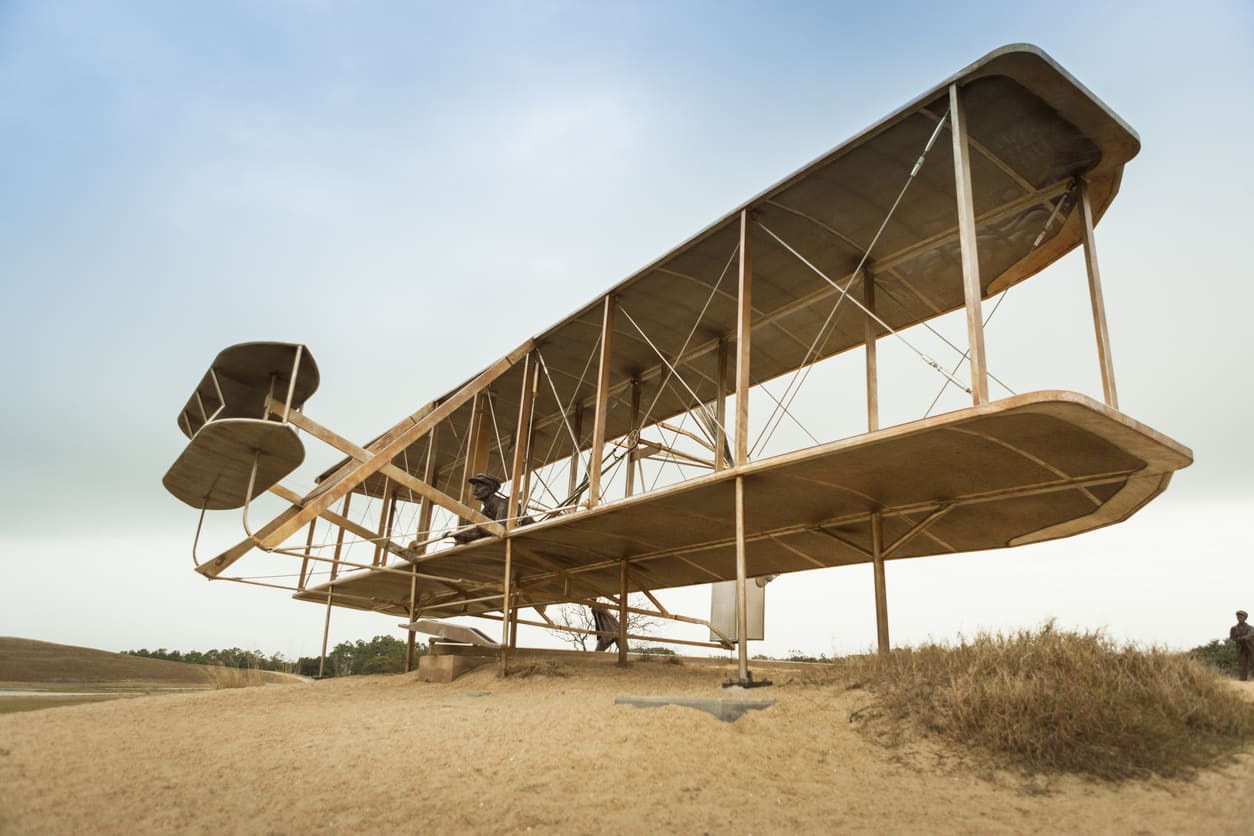 Replica of the Wright brothers' historic airplane, displayed on sandy terrain under a calm sky.