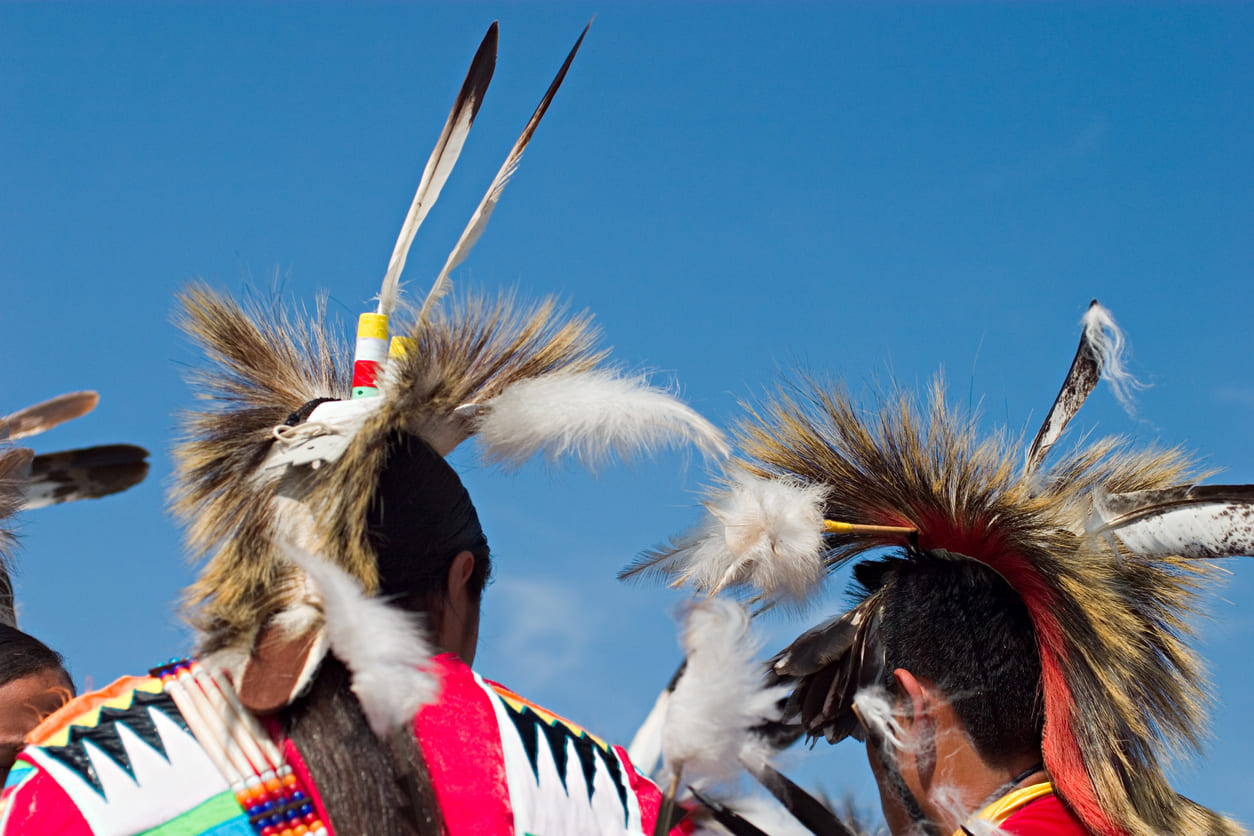 Two individuals wearing traditional Native American headdresses participate in a cultural event against a bright blue sky.