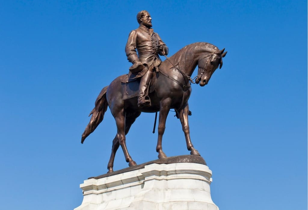 Statue of Robert E. Lee on horseback atop a large pedestal, set against a clear blue sky.