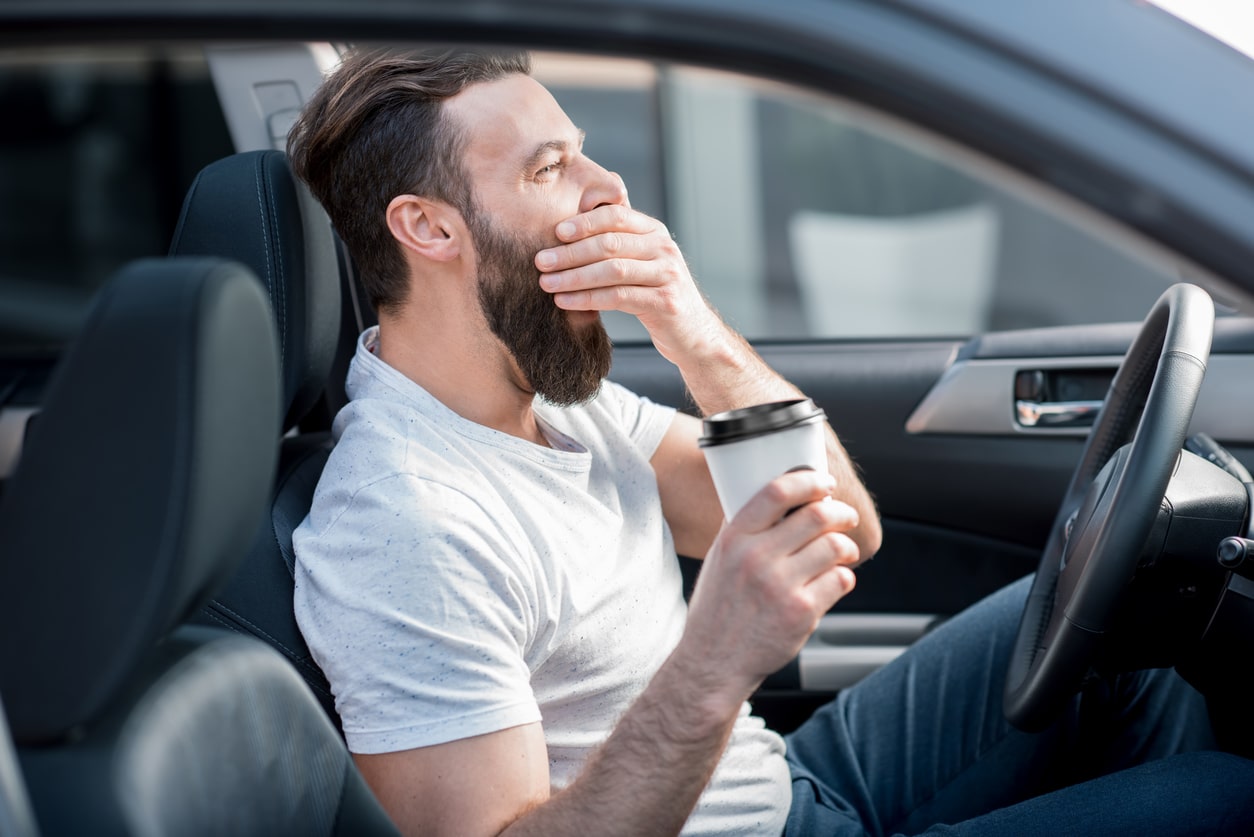 A tired man sits in a car, yawning while holding a takeaway coffee.